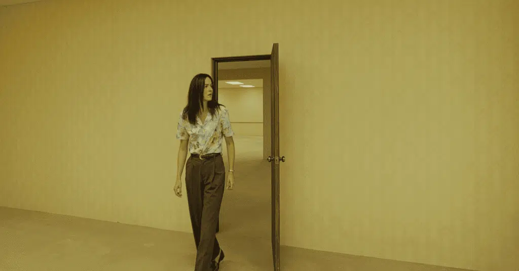 Backrooms: um Não-Lugar, por que o filme da A24 não confia na sua maior força Corredor amarelado dos Backrooms com iluminação fluorescente e personagens perdidos no filme de terror da A24