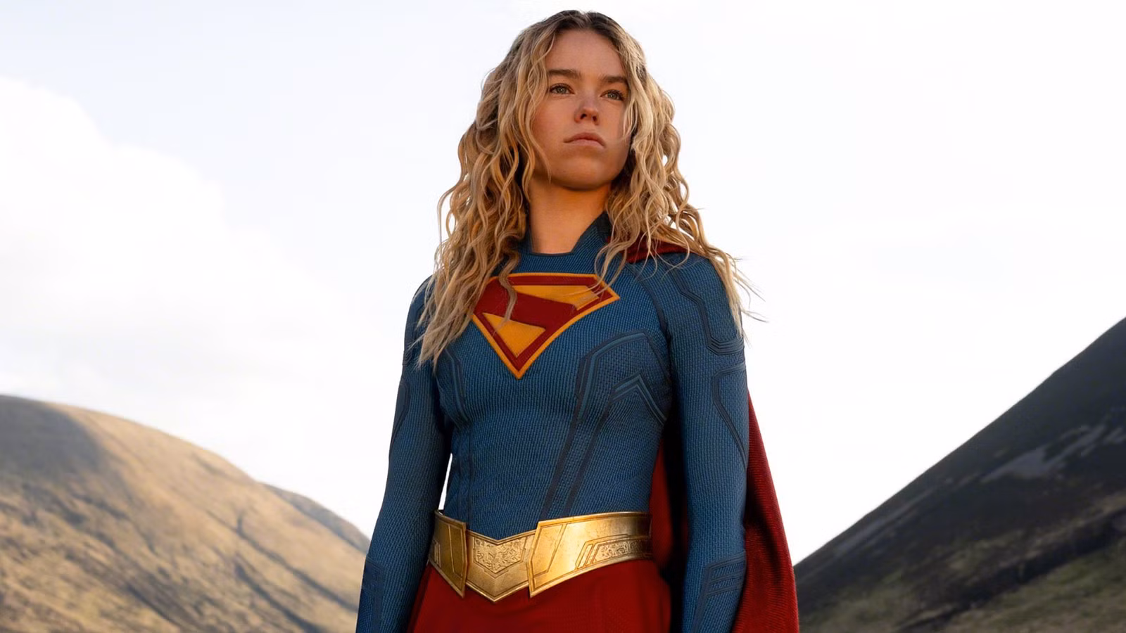 Supergirl em paisagem montanhosa, traje heroico azul e vermelho, olhando para horizonte